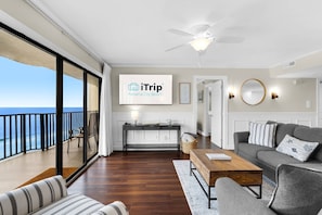 Un vaste espace de vie avec balcon donnant sur l’océan, un ventilateur de plafond et un panneau publicitaire iTrip Panama City Beach fixé au mur.