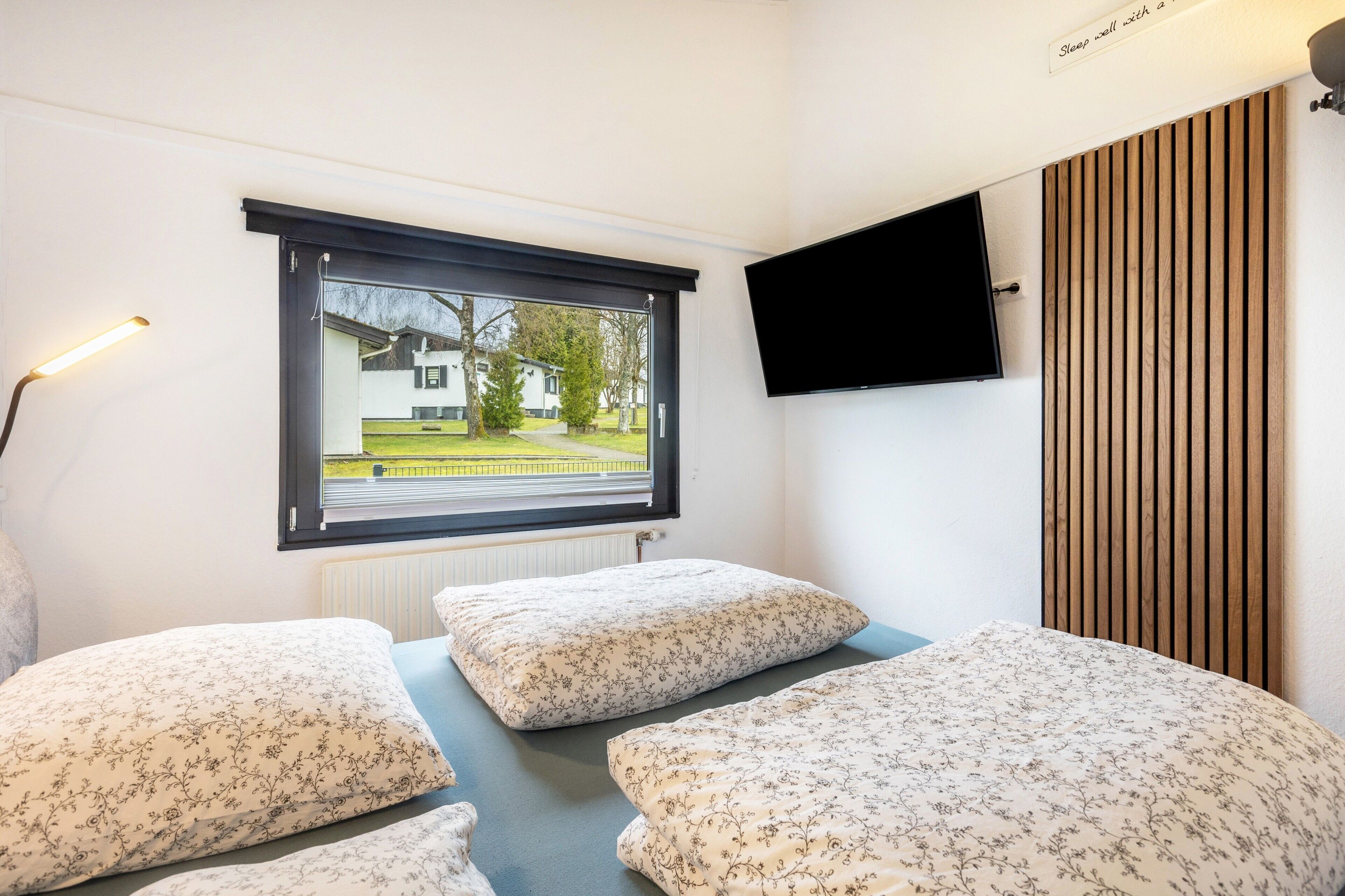 A bedroom with a bed, a window, a wall-mounted TV, and a radiator.