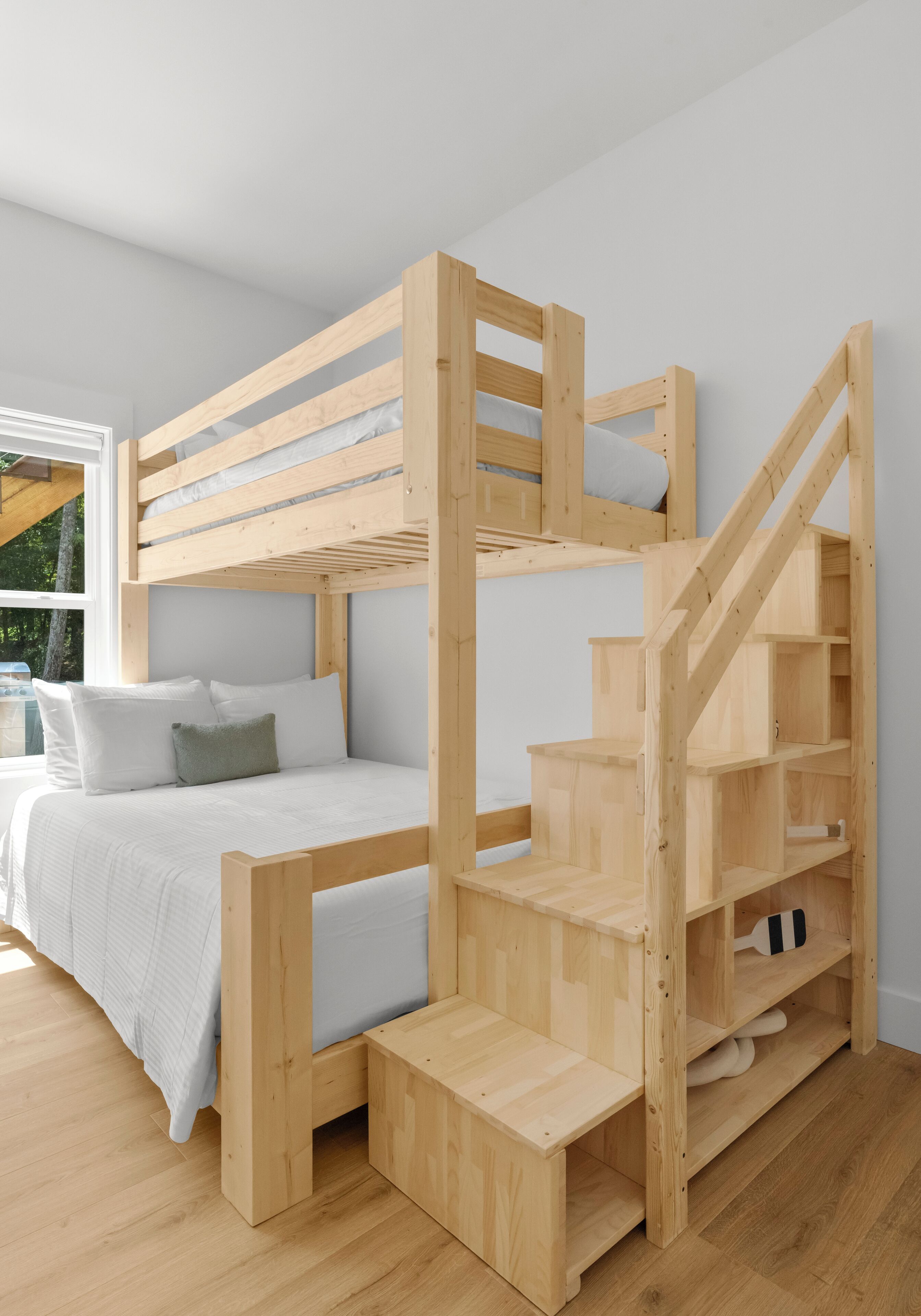 A bunk bed with a wooden ladder and built-in shelves, a bedside table, and a window with a view of trees.