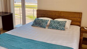 Un dormitorio con una cama grande, cabecera de madera, vistas a la playa y una mesita junto a la ventana.