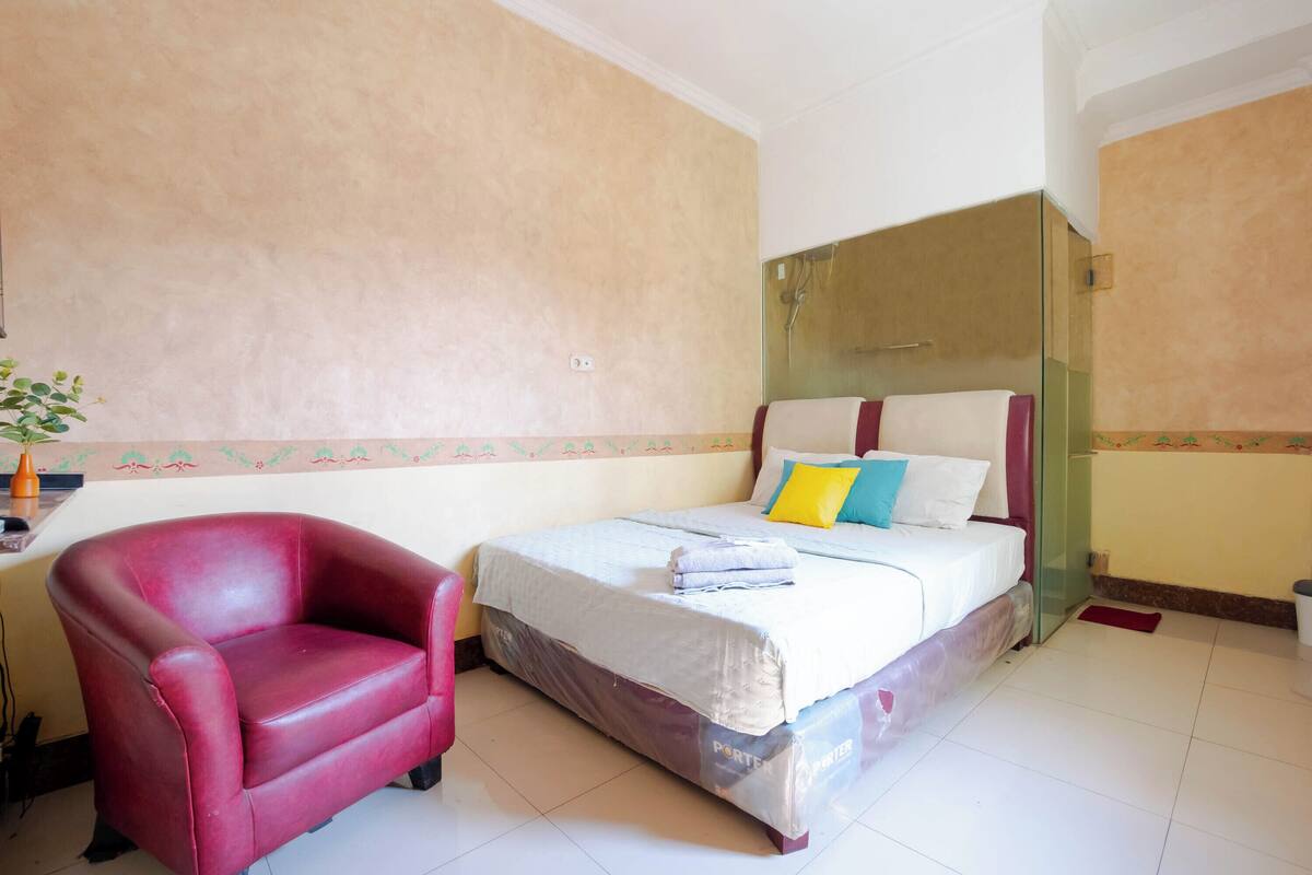 A hotel room with a bed, a red armchair, a small table with a vase, and a wall with a decorative border.