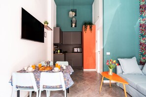 Cozinha moderna com televisão de tela plana, área de jantar com mesa posta para o café da manhã e sala de estar com sofá e um vaso de flores.