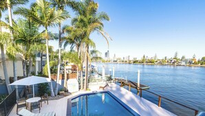 A pool area with lounge chairs and umbrellas, surrounded by palm trees and overlooking a body of water with a city skyline in the background.