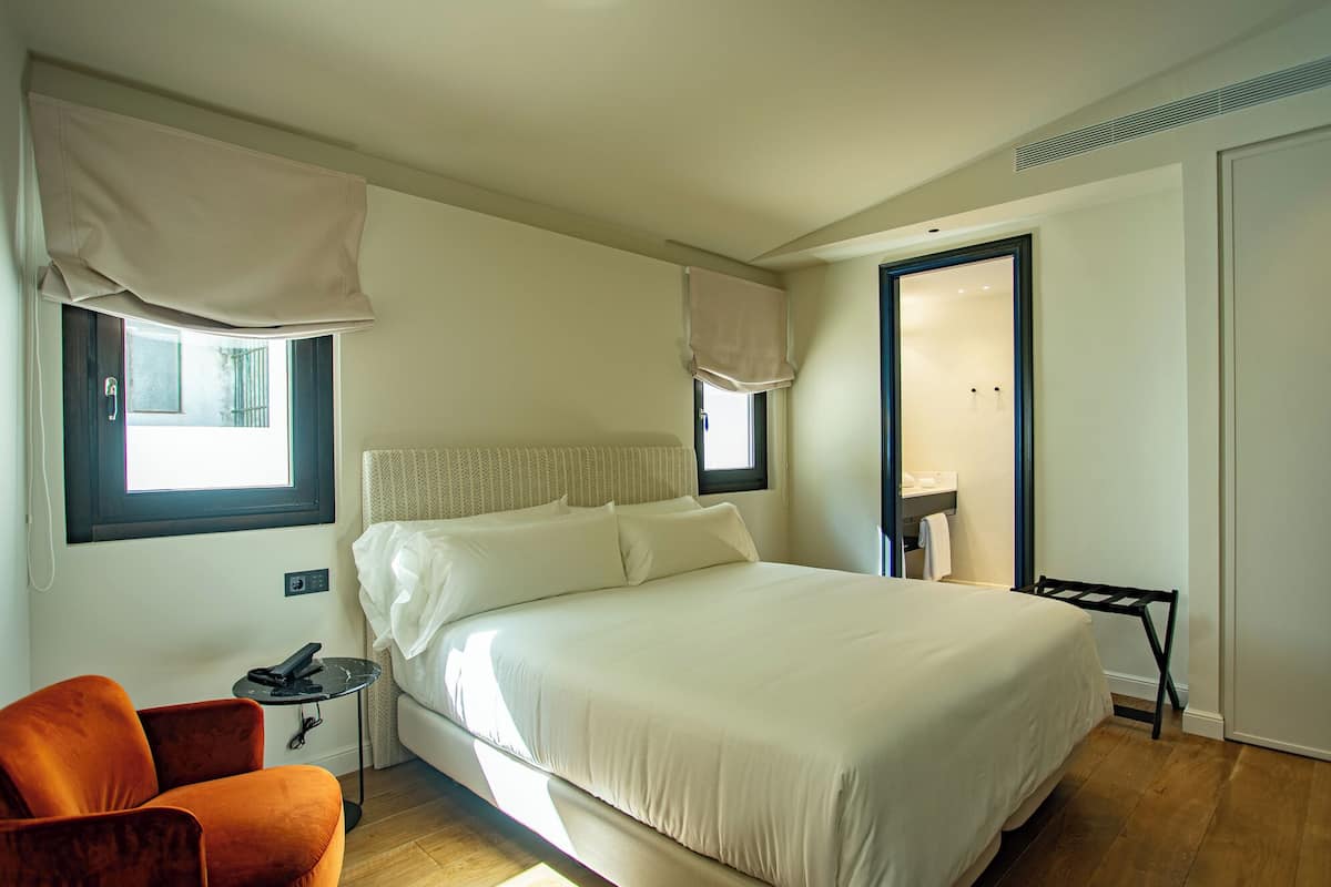 Un dormitorio con cama, una silla, mesita de noche y ventana con cortinas.