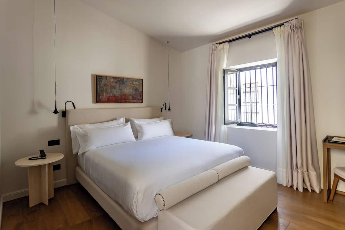 Un dormitorio ordenado con una cama grande, una mesita de noche, una pequeña mesa redonda y una ventana con cortinas.