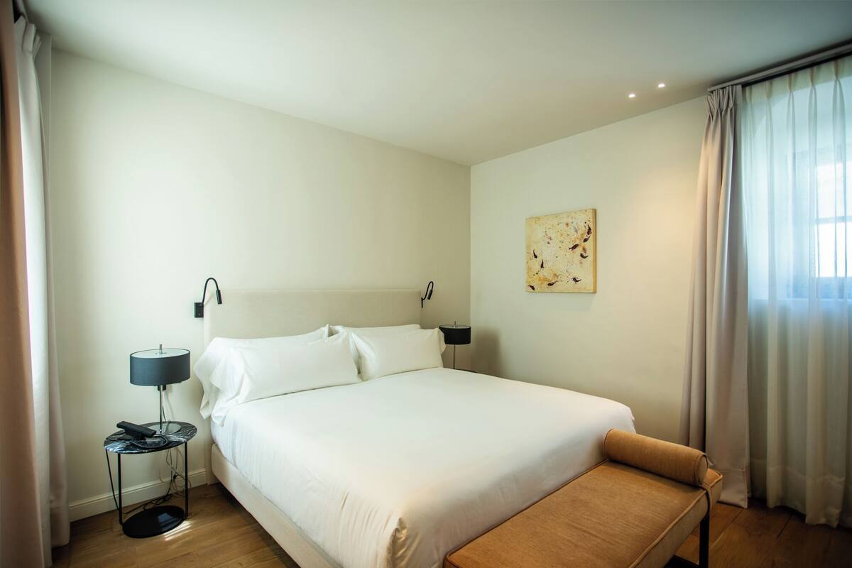 Habitación de hotel con una cama grande, una mesita de noche con lámpara, una mesita auxiliar pequeña y un banco.