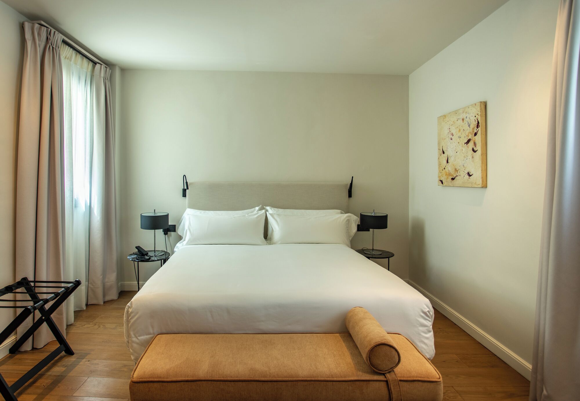 A hotel room with a large bed, a bedside table with a lamp, a small table with a phone, a bench, and a painting on the wall.