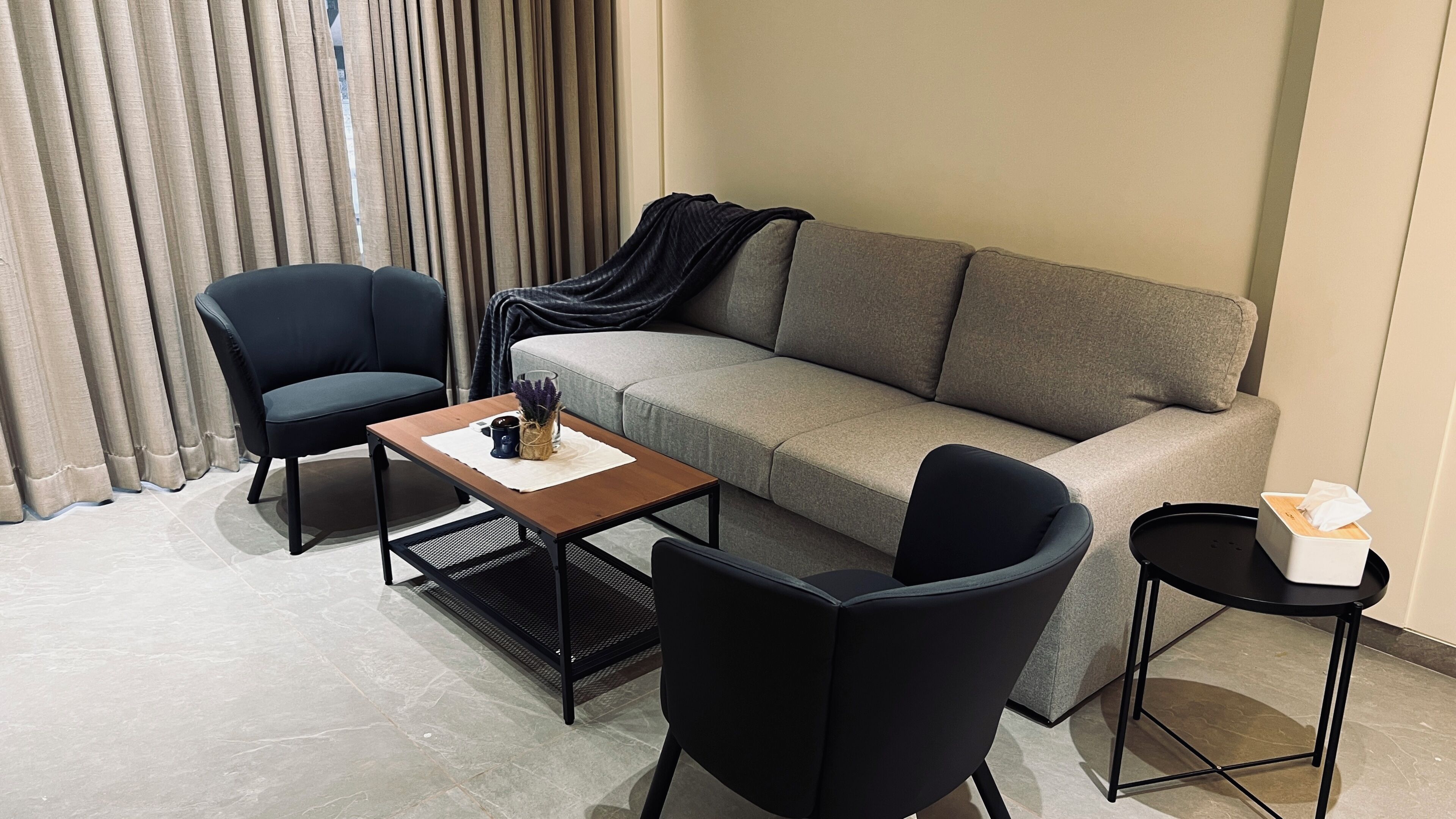 Una sala de estar moderna con un sofá gris, dos sillas negras, una mesa de centro de madera y una mesita auxiliar negra con una caja de pañuelos.