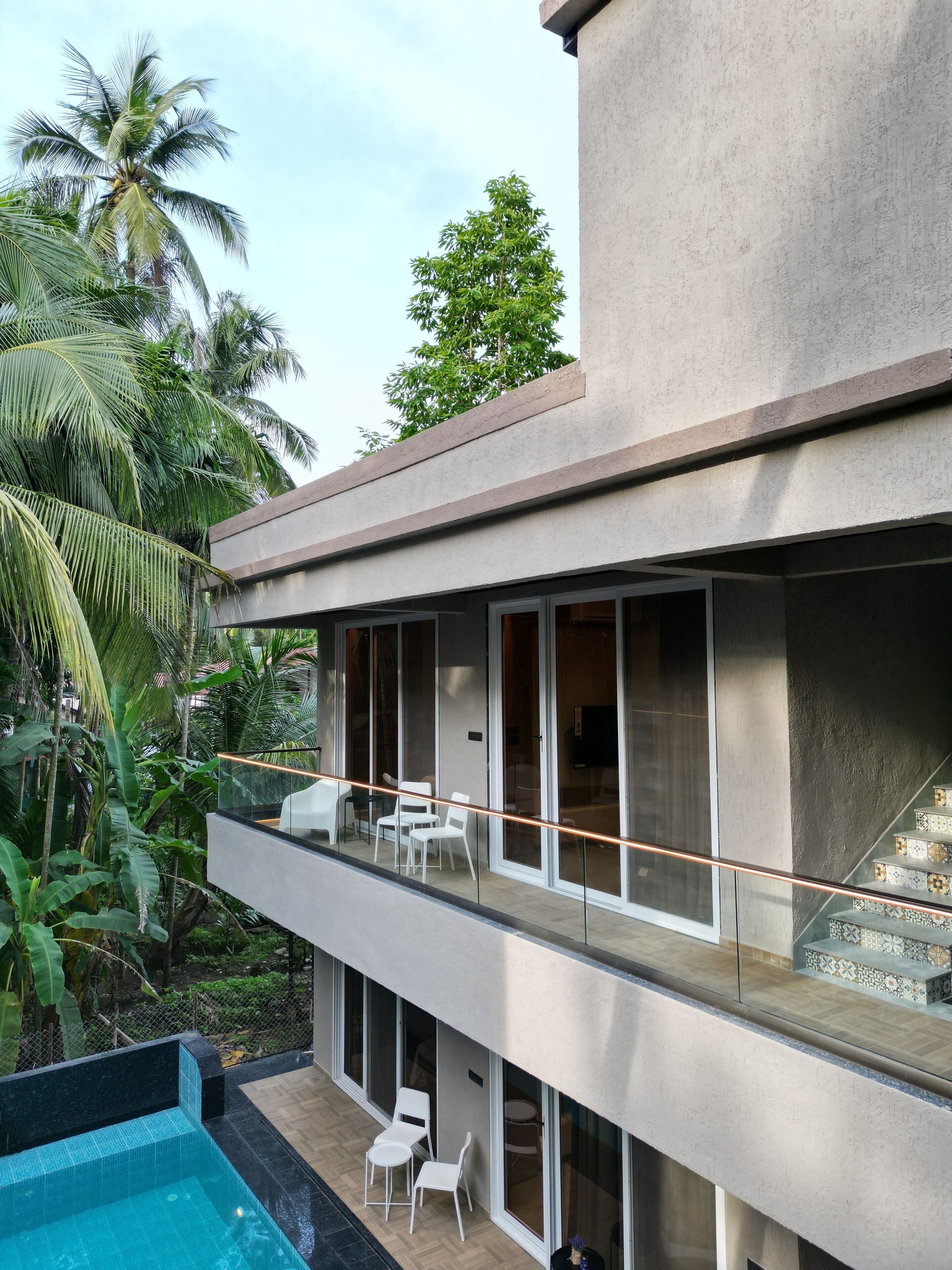 Un bâtiment moderne à deux étages, doté d’un balcon en verre, d’une piscine et d’une végétation tropicale luxuriante.