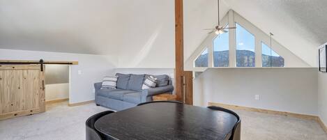 A spacious loft with a high ceiling, a dining table, a sofa, and large windows offering a view of the mountains.