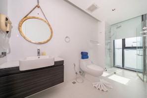 A modern bathroom with a white sink, dark vanity, a round mirror with a gold frame, a toilet, and a glass-enclosed shower area. - Tropical Suite A602 at Viva Patong Condo (Patong)
