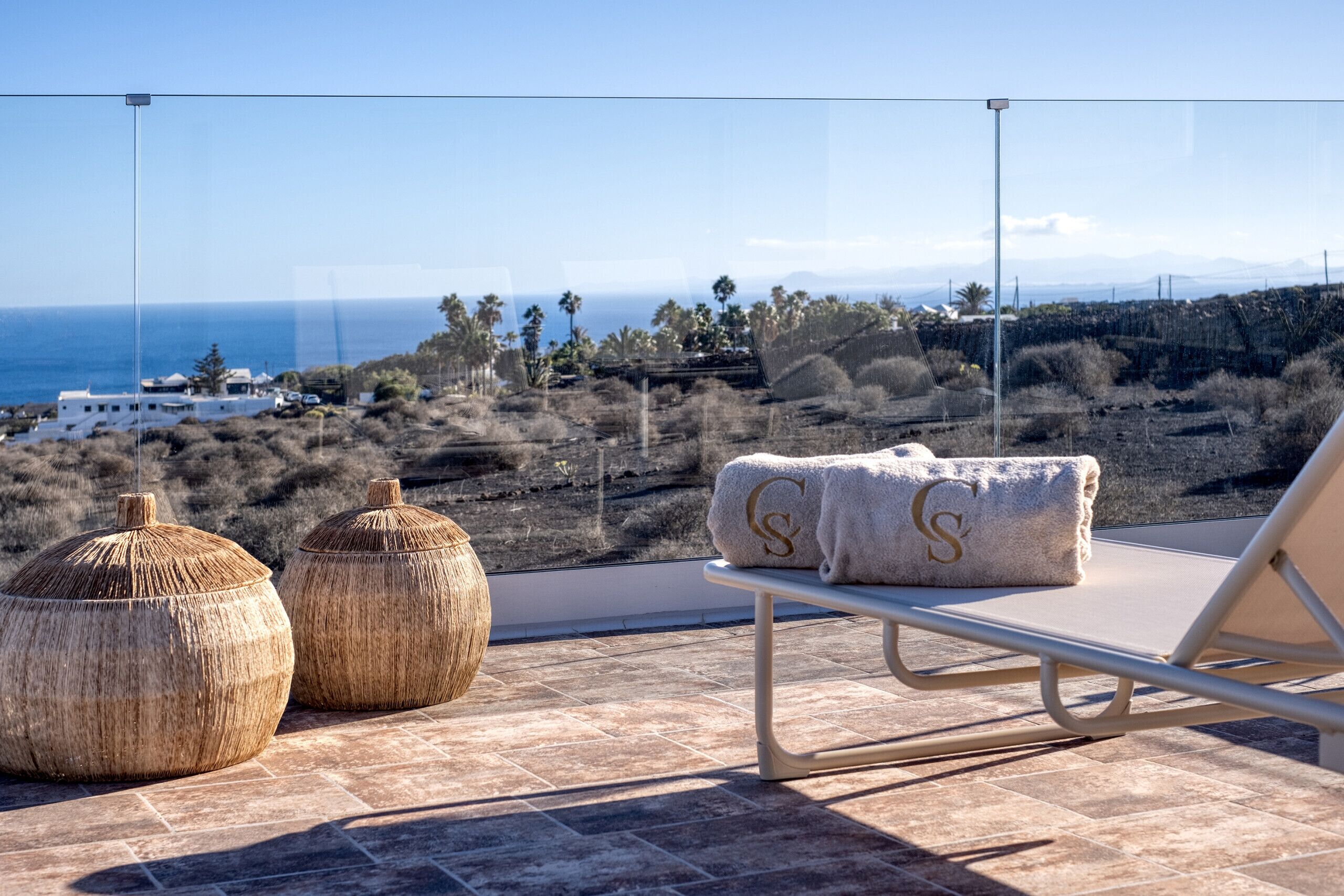 Een zonnig terras met een ligstoel en twee geweven poufs, met uitzicht op een kustlandschap met palmbomen en een verre zee.