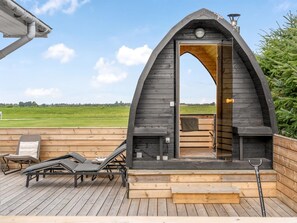 Une cabane en bois dotée d’une entrée en arc de cercle, d’une terrasse avec des chaises longues et d’une vue sur un champ verdoyant.