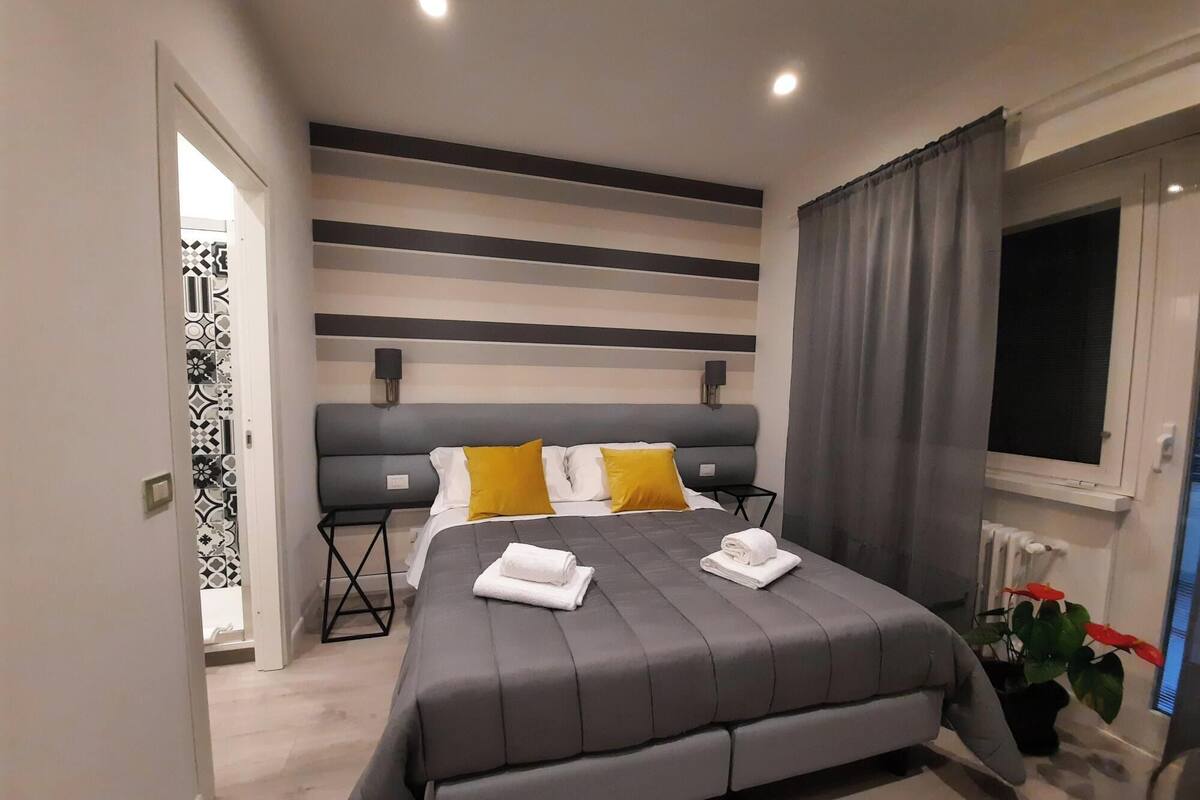 Ein modernes Schlafzimmer mit einem großen Bett, grauer Bettwäsche, einem grauen Kopfteil und einem Fenster mit Vorhängen.