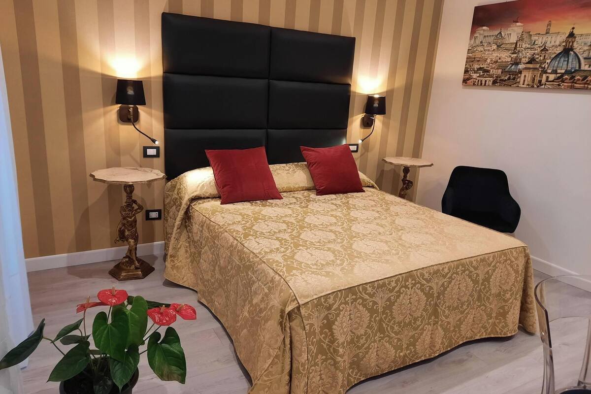 Ein ordentlich eingerichtetes Schlafzimmer mit einem großen Bett, zwei Nachttischlampen, einem kleinen Tisch, einem Sessel und einer Topfpflanze.