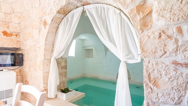 A stone-walled room with a clear blue pool, white curtains, and a white chair. - 4152 Trulli Salsapariglia by Perle di Puglia (Putignano)