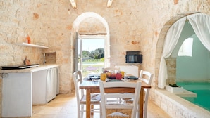 A rustic stone interior with a dining table, chairs, a small kitchen area, and a view of a swimming pool. - 4152 Trulli Salsapariglia by Perle di Puglia (Putignano)