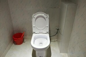 Des toilettes avec un abattant fermé, un seau rouge et une tuyauterie apparente.