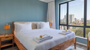 A neatly made bed with white linens and a folded towel, a wooden nightstand with a lamp, and a large window offering a city view. - Alba Gardens 1Bed by YourHost  (Nairobi)