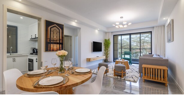 A modern dining area with a wooden table set for four, a television mounted on the wall, and a view of the outdoors through large windows. - Alba Gardens 1Bed by YourHost  (Nairobi)