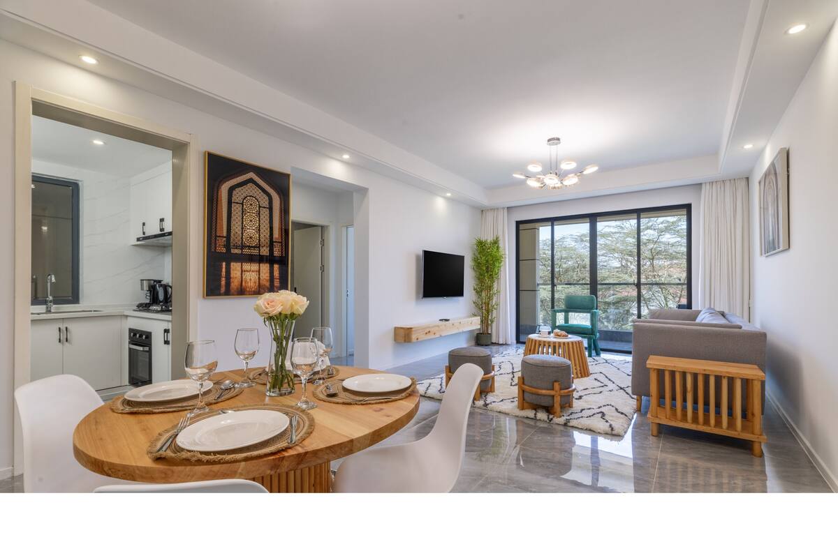 A modern dining area with a wooden table set for four, a television mounted on the wall, and a view of the outdoors through large windows.