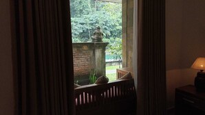 Una habitación con vista a un jardín a través de una ventana, un banco de madera y una lámpara en una mesita de noche.