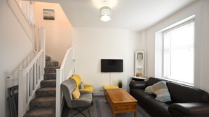 Una sala de estar con una escalera, un televisor de pantalla plana, una mesa de centro de madera, un sofá de cuero negro y un ventanal grande.