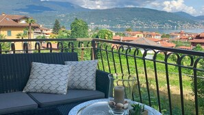 A balcony with a wicker sofa, two patterned cushions, a glass table with a candle and seashells, and a scenic view of a lake and mountains. - Apartment in Baveno with Lake Maggiore Views (Baveno)