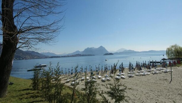 A beach with rows of umbrellas and lounge chairs, a boat on the water, and mountains in the background. - Apartment in Baveno with Lake Maggiore Views (Baveno)