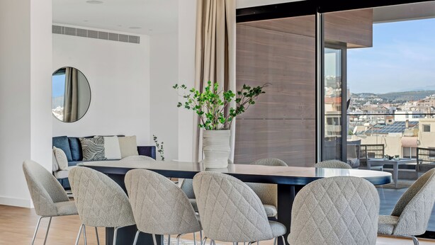 A modern dining area with a large table, grey upholstered chairs, a potted plant, and a view of the city through large windows. - Sanders Crystal 2 Limassol (Limassol)