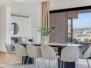 A modern dining area with a large table, grey upholstered chairs, a potted plant, and a view of the city through large windows. - Sanders Crystal 2 Limassol (Limassol)