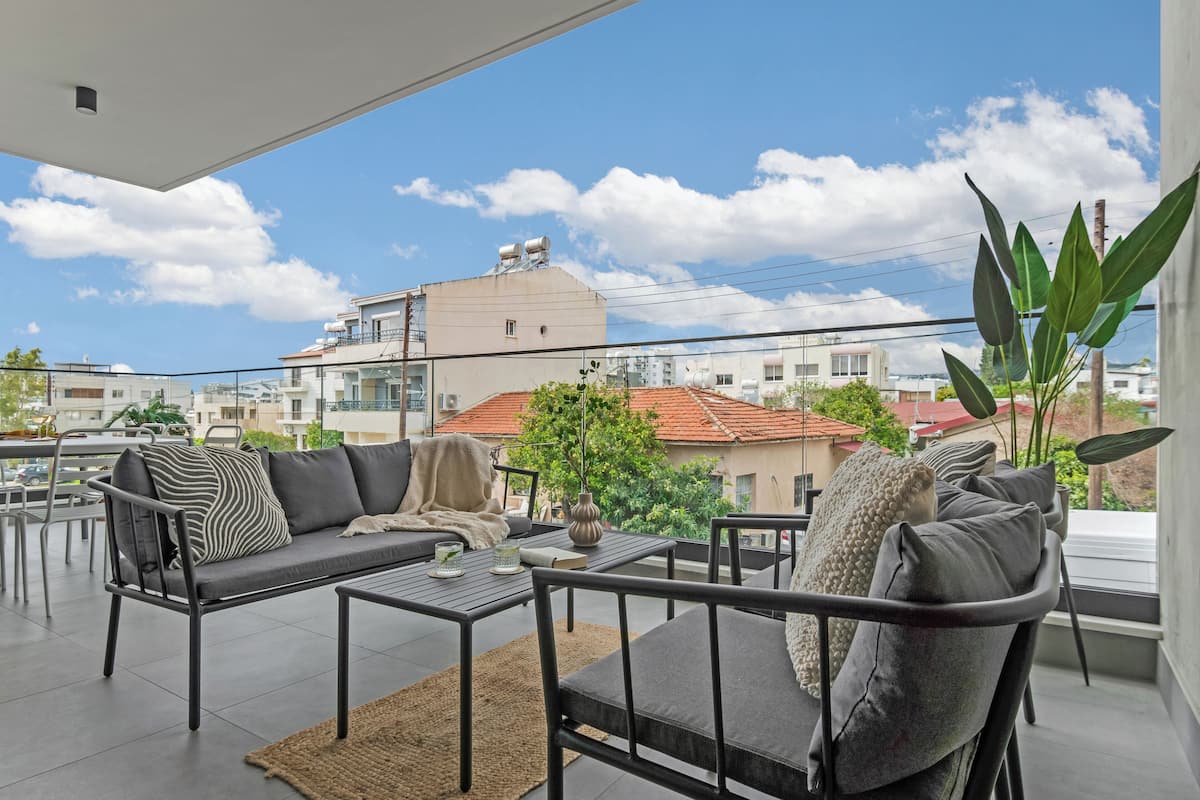 A balcony with a sofa, chairs, and a table, overlooking a cityscape with buildings and a clear sky.