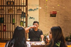 A traditional tea room with a table set for a meal, a tea station with various tea containers, and a decorative wall with a mountainous landscape. - Jibolong Hotel (Lijiang)