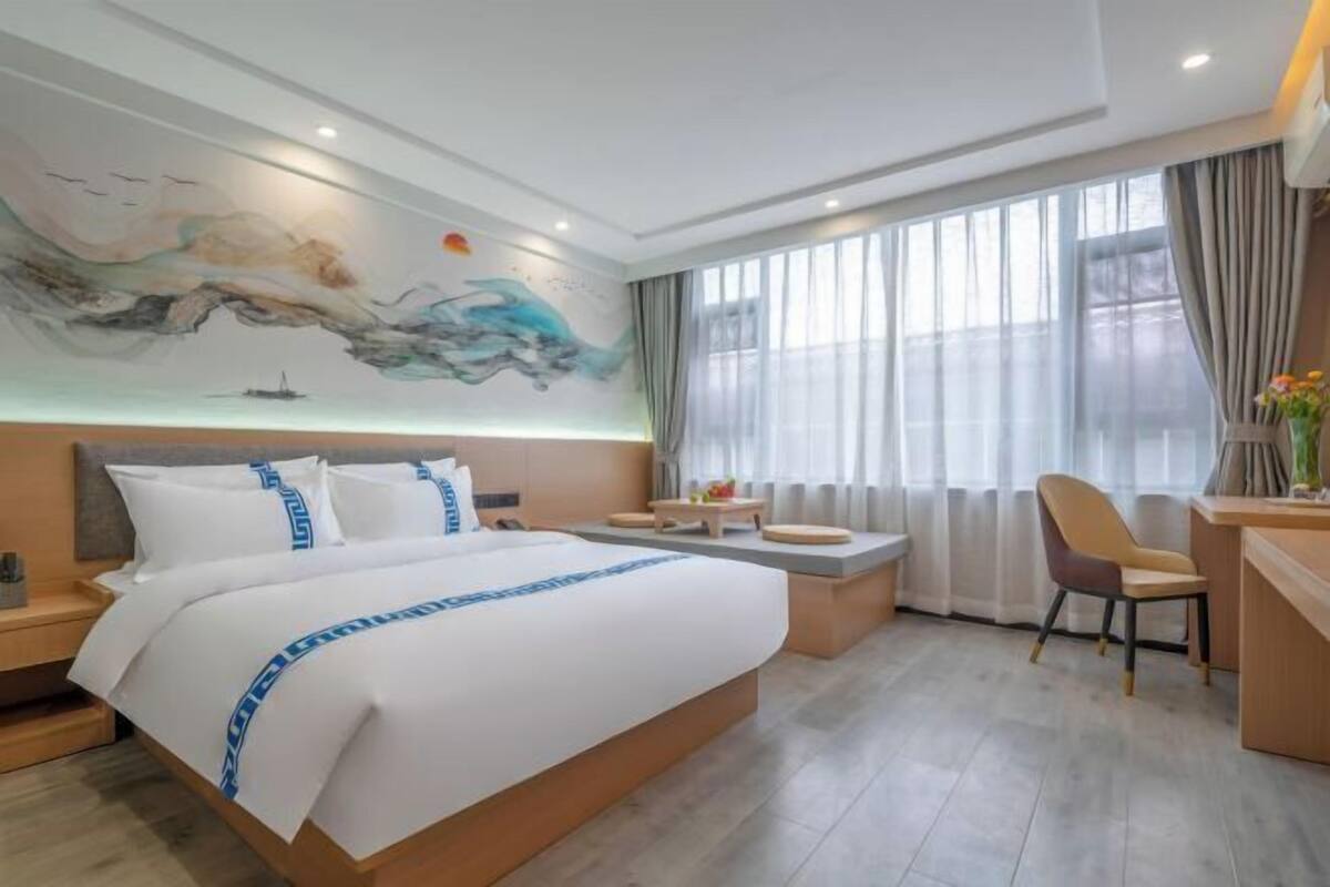 A modern hotel room with a large bed, a desk, a chair, and a mural on the wall.