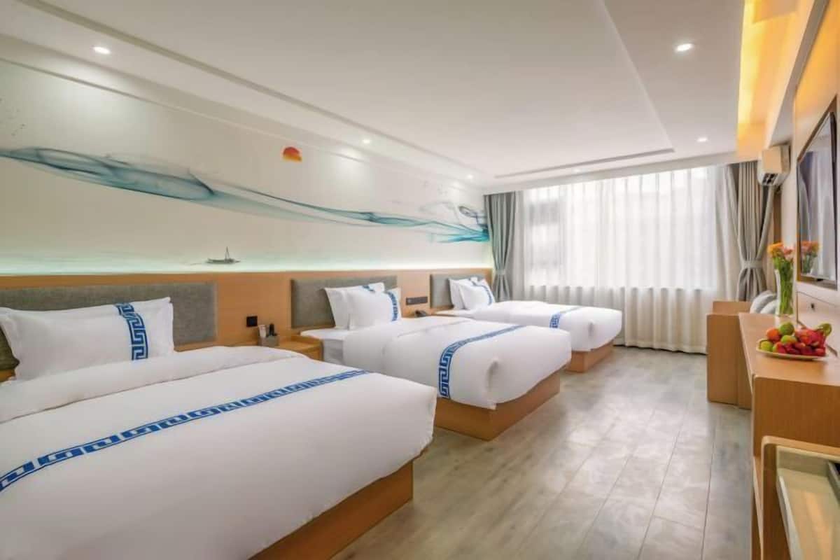 A hotel room with three beds, a large window with curtains, a desk with a fruit bowl, and a modern abstract wall design.