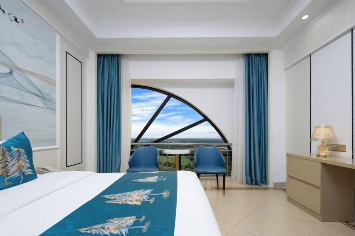 Une chambre d’hôtel avec un grand lit, deux fauteuils, un bureau et une vue dégagée sur l’extérieur grâce à une grande fenêtre.