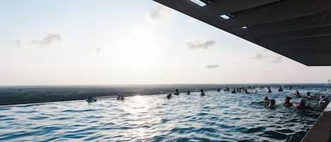Une piscine extérieure où des personnes nagent, située sous un pont, avec un ciel dégagé.
