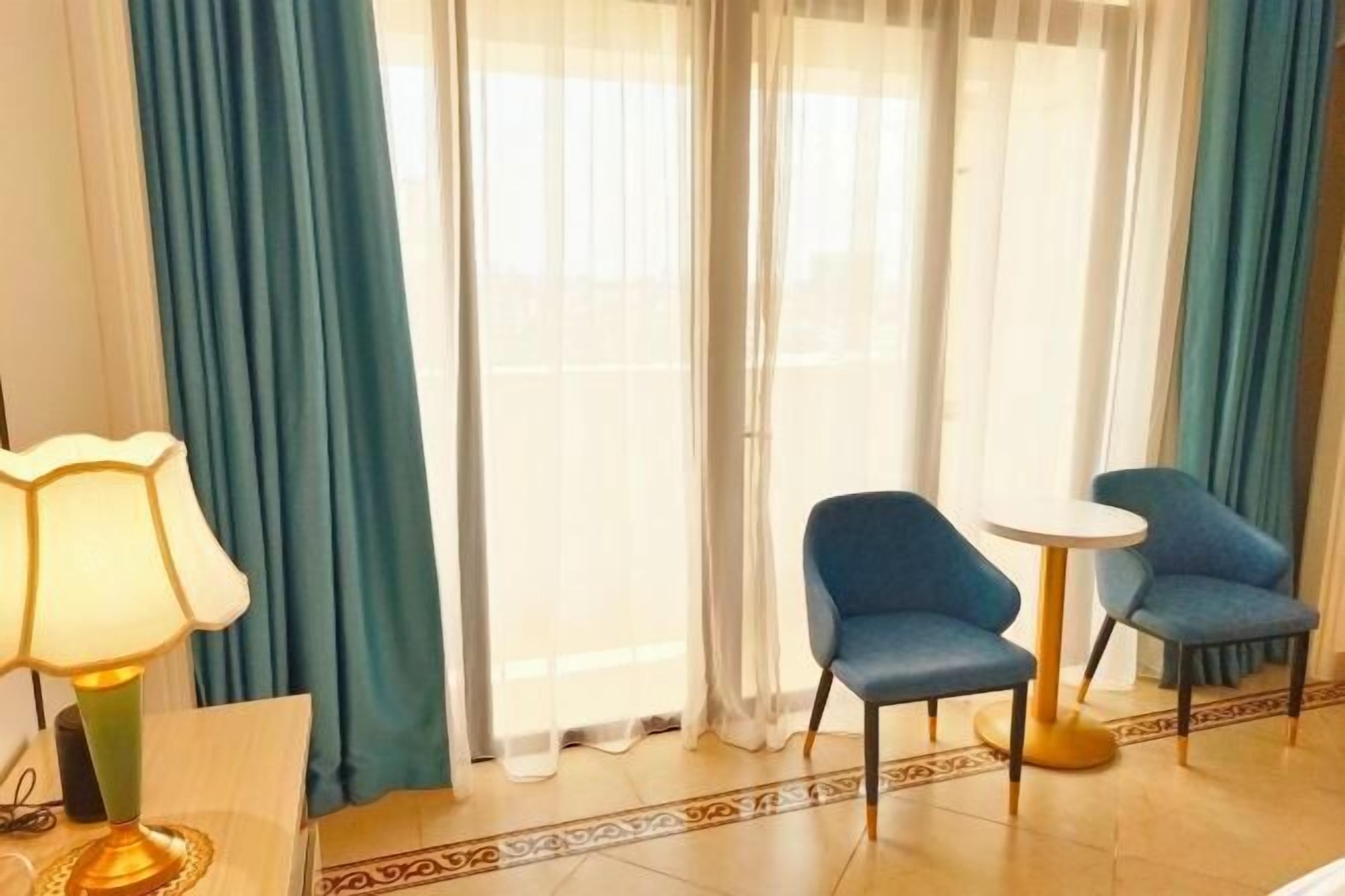 Une chambre d’hôtel avec un lit, une table avec une lampe, deux chaises bleues, une petite table ronde et une fenêtre avec des rideaux bleus.