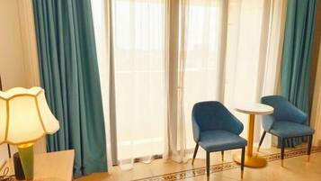 Une chambre d’hôtel avec un lit, une table avec une lampe, deux chaises bleues, une petite table ronde et une fenêtre avec des rideaux bleus.