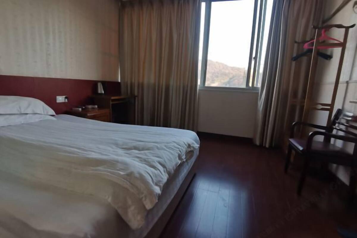 A hotel room with a bed, a nightstand, a chair, a window with curtains, and a coat rack.
