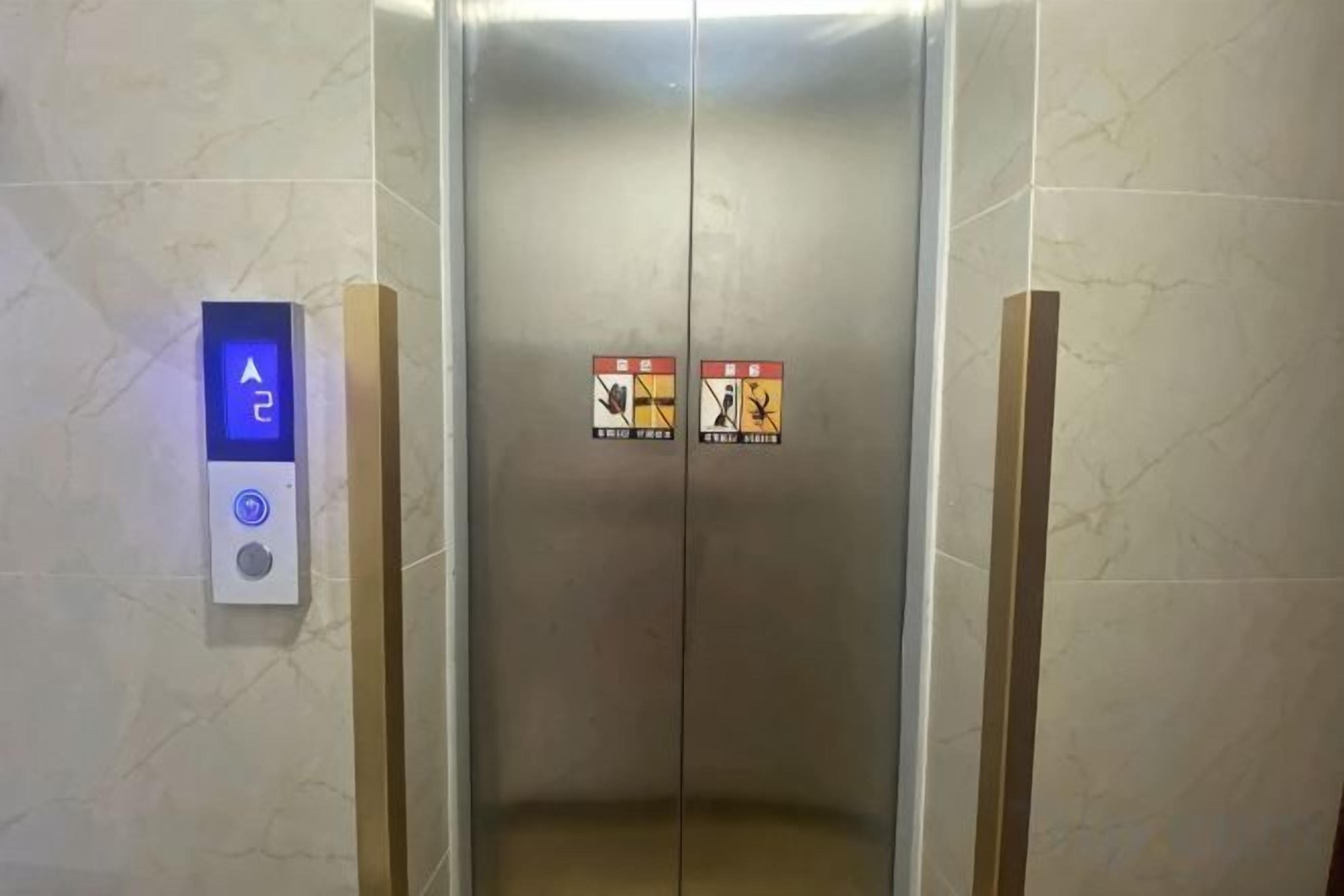 Un ascensor con un panel de control que muestra una flecha hacia arriba y un botón, flanqueado por dos letreros de advertencia de seguridad.