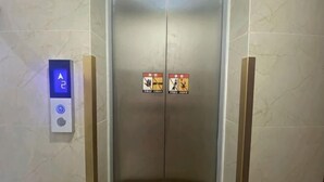 Un ascensor con un panel de control que muestra una flecha hacia arriba, dos señales de advertencia de seguridad y una pared de mármol.
