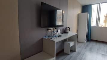 A modern hotel room with a flat-screen TV mounted on the wall, a white desk with a kettle, and a refrigerator.