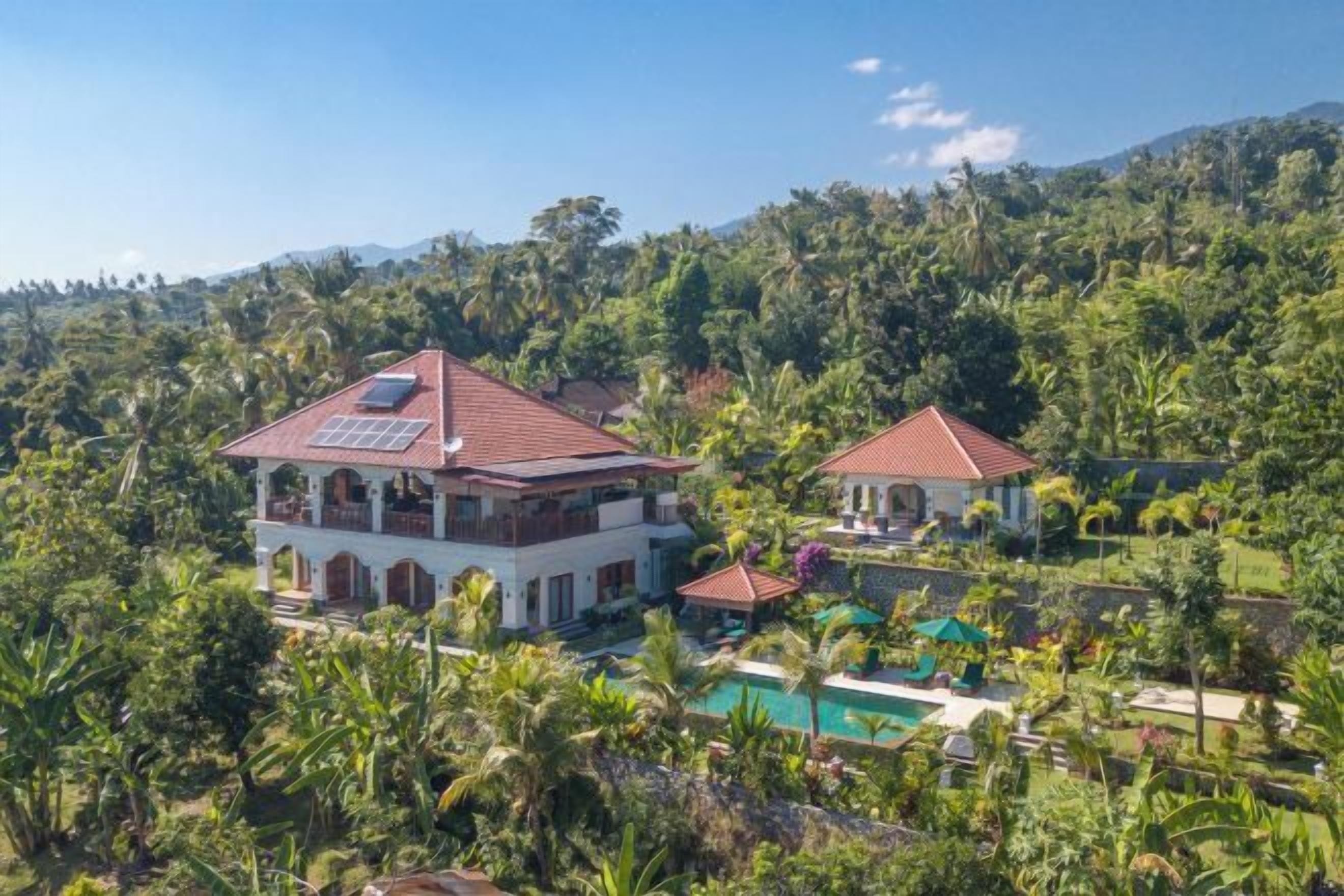 Une maison à deux étages avec une piscine, entourée d’une végétation luxuriante et avec des collines en arrière-plan.