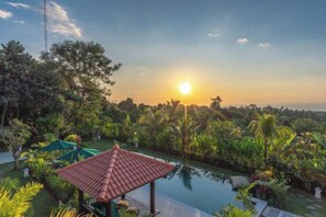 A pool with a red-roofed pavilion, surrounded by lush greenery and a sunset in the background. - Villa Mata Hari (Bali)