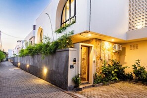 A modern building with a paved walkway, a stone wall with greenery, and a well-lit entrance. - Floro Villa Canggu by Dhananjaya Hospitality (Munggu)