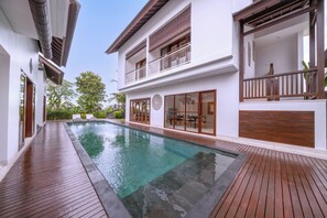 A modern pool area with wooden decking, a clear pool, and a two-story building with large windows and a balcony. - Floro Villa Canggu by Dhananjaya Hospitality (Munggu)