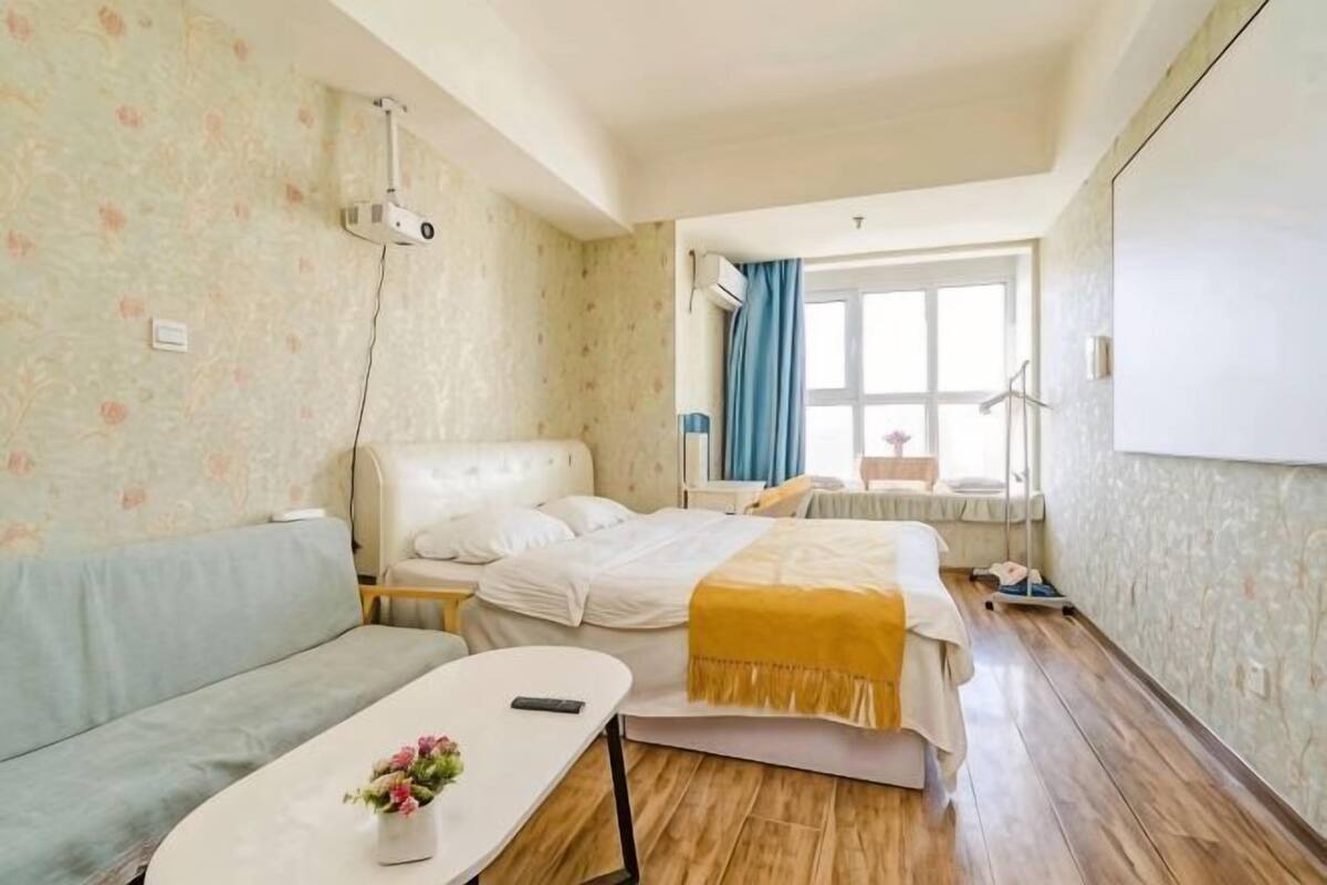 A hotel room with a bed, a sofa, a small table with a flower, a projector, and a window with curtains.