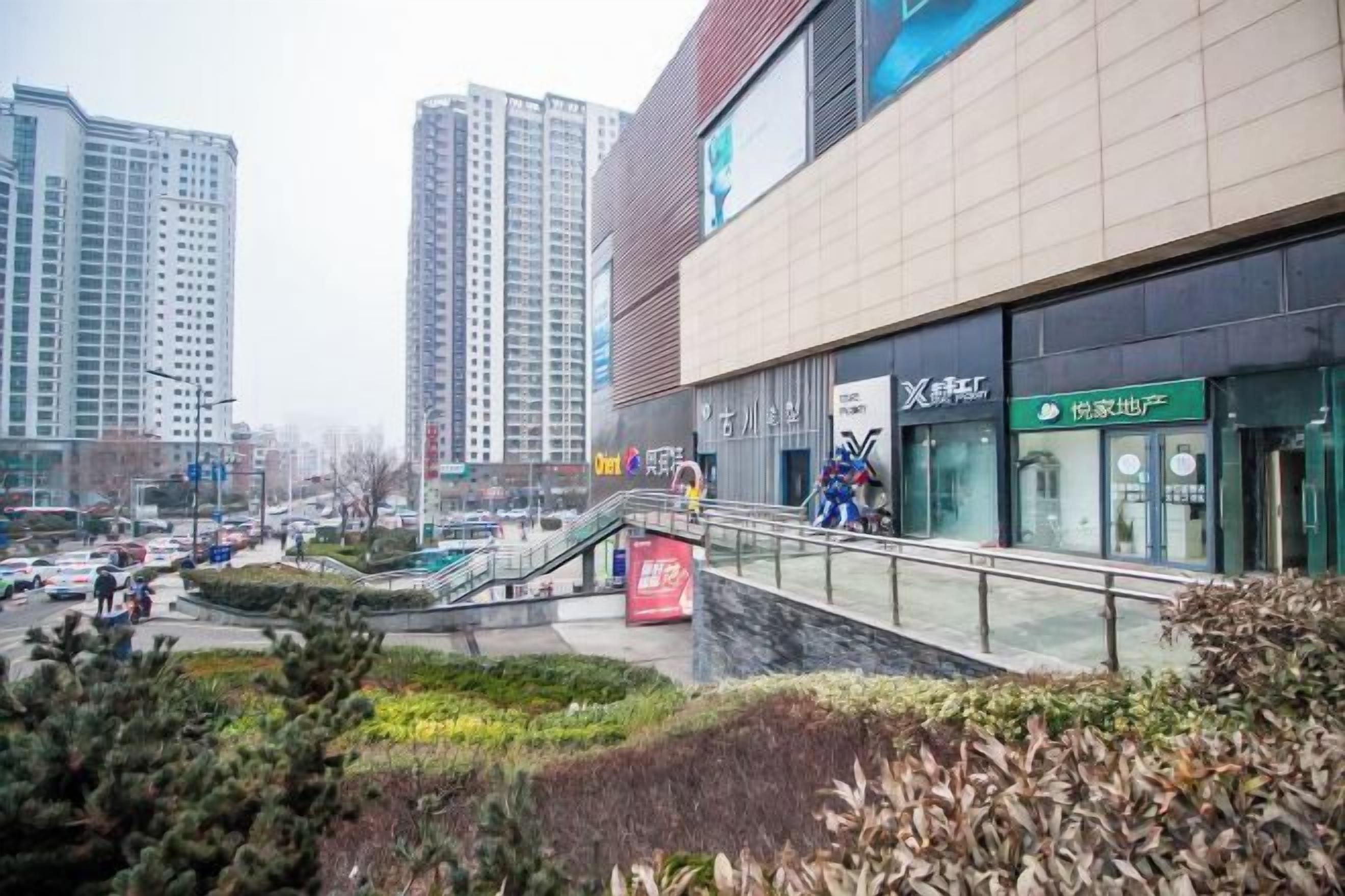 A modern building with a glass facade, a pedestrian walkway, and a landscaped area with shrubs.