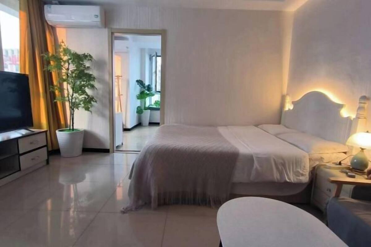 A hotel room with a bed, bedside table, lamp, TV, potted plant, and a view of another room.